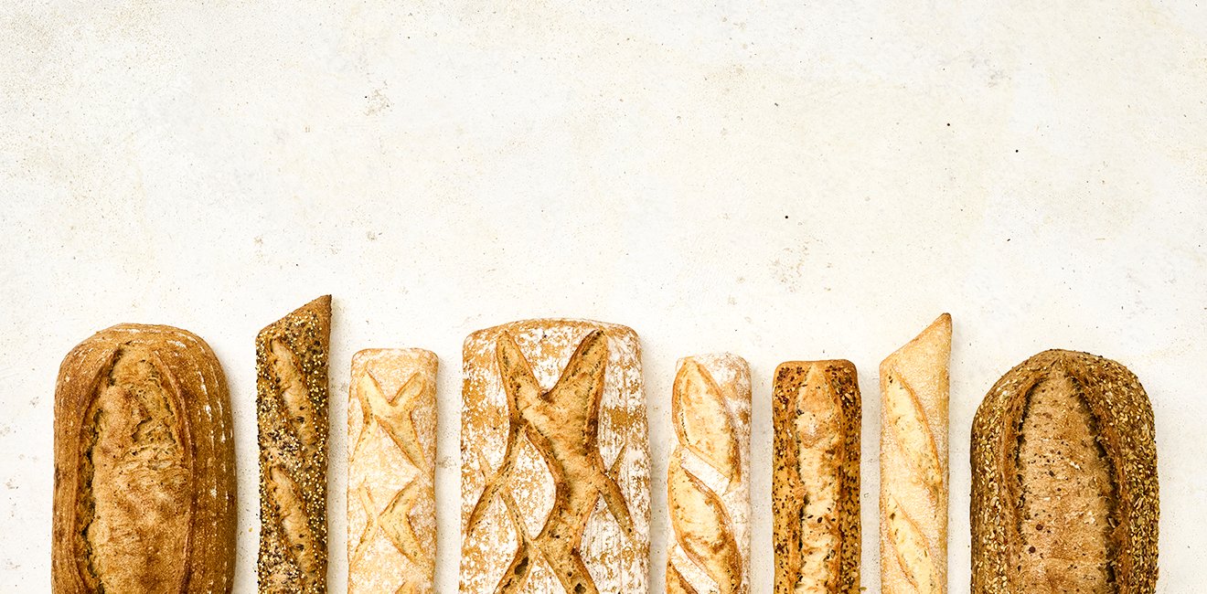 Différents pains alignés : pain rustique, baguette aux graines, pain blanc à croûte farinée, pain polka, baguette dorée, baguette aux céréales et pain aux graines.